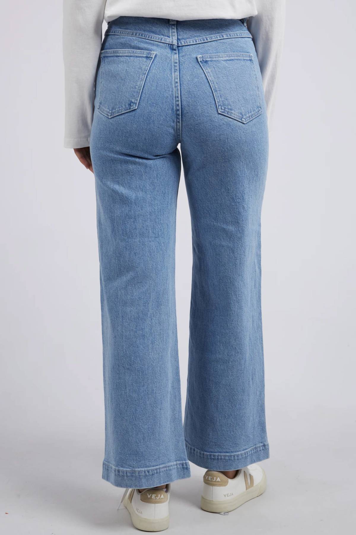 Person wearing light blue Foxwood Sarah Wide Leg Jean, featuring a high-rise, rigid denim and wide leg cut, paired with white Veja sneakers, shown from the back against a plain light background.