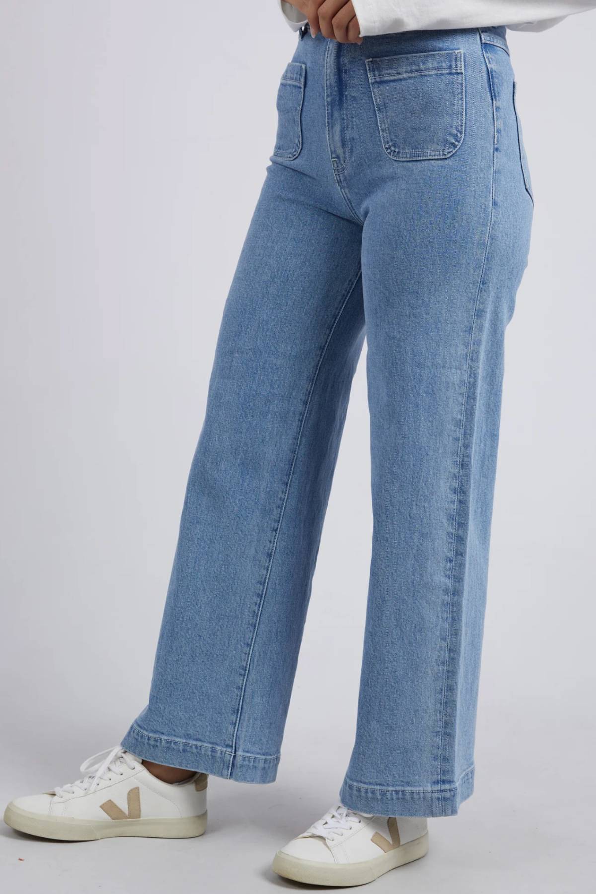 Wearing Foxwood Sarah Wide Leg Jeans crafted from rigid denim, paired with a white long-sleeve top and white sneakers, a person stands against a plain light background with only the lower half of their body visible.