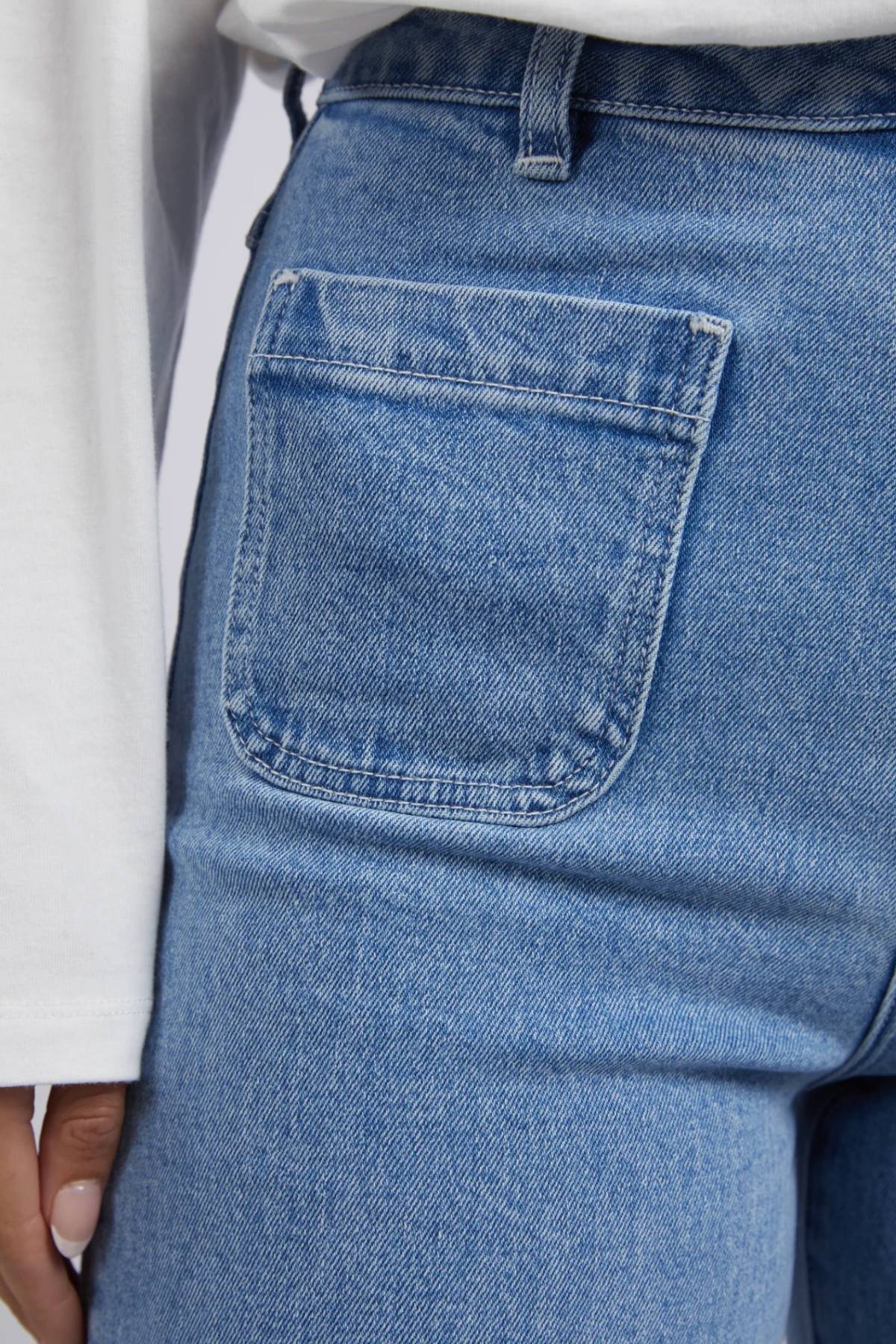Close-up of a person wearing Foxwood Sarah Wide Leg Jean in light blue rigid denim with a high-rise fit and a white long-sleeve shirt, focusing on the back pocket and the person's hand by their side.