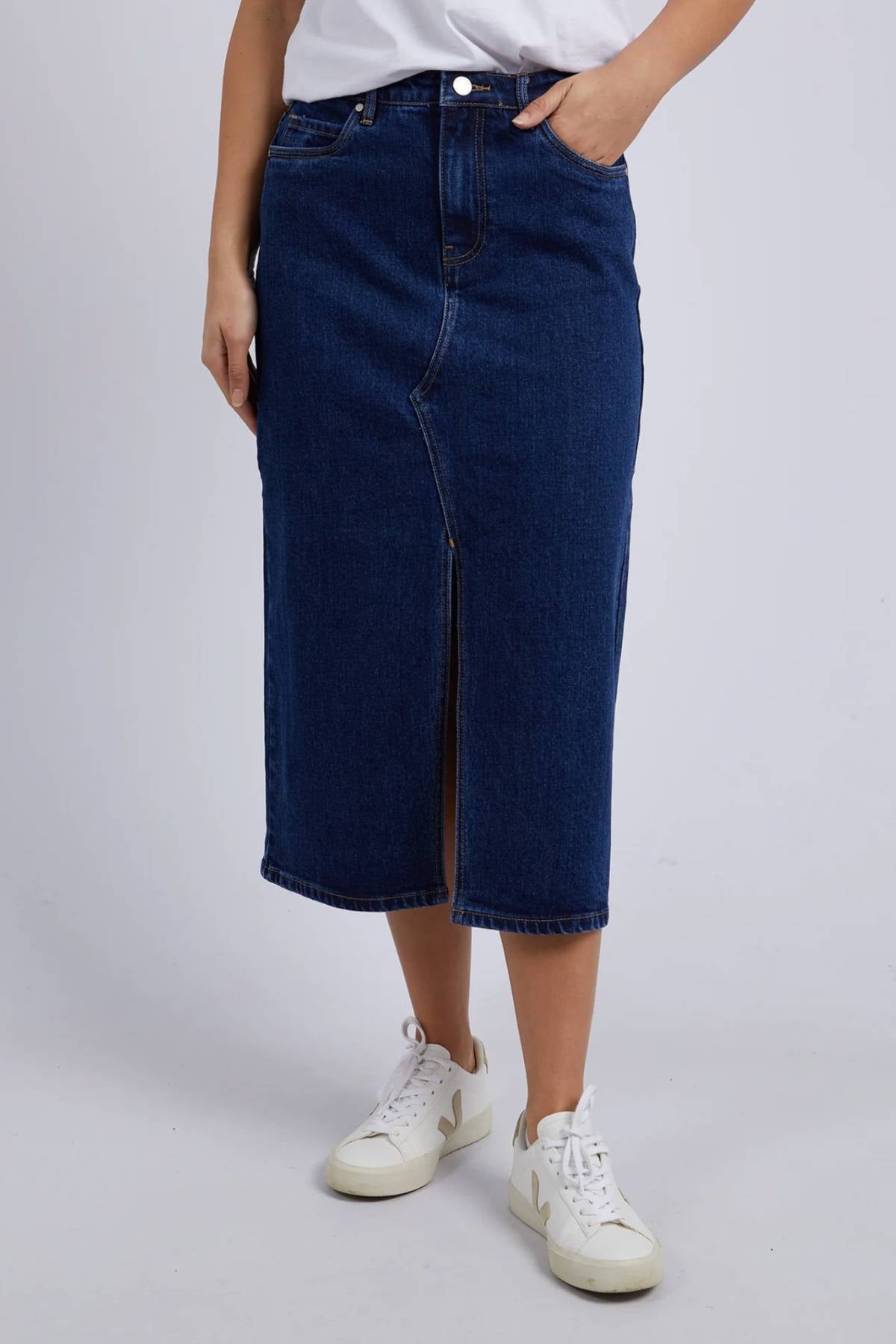 A person wearing a white t-shirt, Foxwood Scout Midi Skirt in dark blue stretch denim with a front slit, and white sneakers stands against a plain light background.
