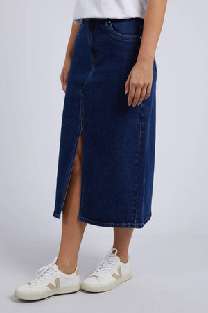 A person wears a white t-shirt, the Foxwood Scout Midi Skirt in dark blue stretch denim with a front slit, and white sneakers with beige accents, standing against a plain light grey background.