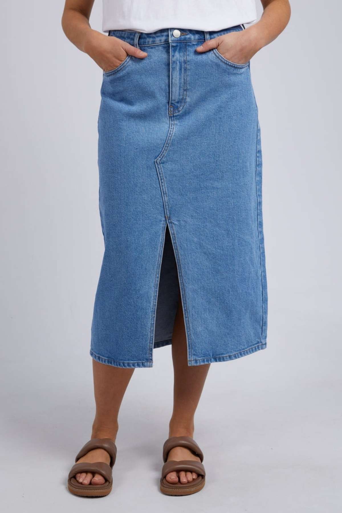 A person models the chic Foxwood Scout Midi Skirt in blue denim featuring a front slit, paired with a white top and brown sandals. Standing stylishly against a neutral background, they casually place their hands in the skirt's pockets.