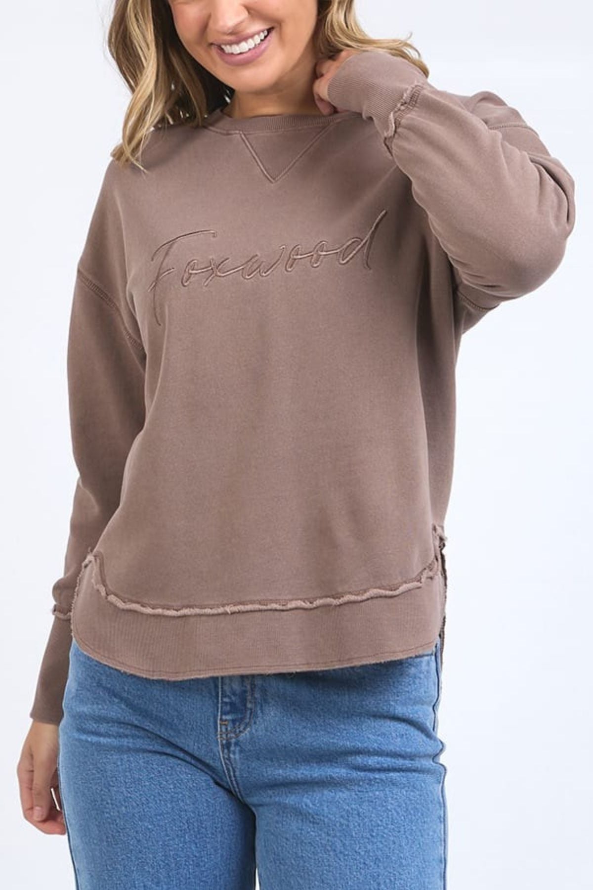 A woman in the Foxwood Signature Crew—a 100% cotton crewneck sweater with "Foxwood" embroidery—pairs it with blue jeans, smiling as she touches her hair against a plain light background.