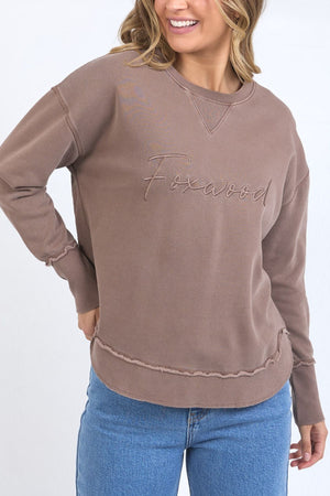 A woman wears the Foxwood Signature Crew, a 100% cotton crewneck with "Foxwood" embroidered on the front, paired with blue jeans. She smiles confidently and poses with one hand on her hip.