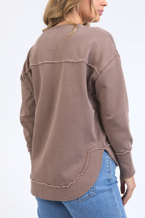 A woman with blonde hair wears the Foxwood Signature Crew, a 100% cotton sweater with raw edge seams, paired with light blue jeans. She stands facing away against a plain, light background.