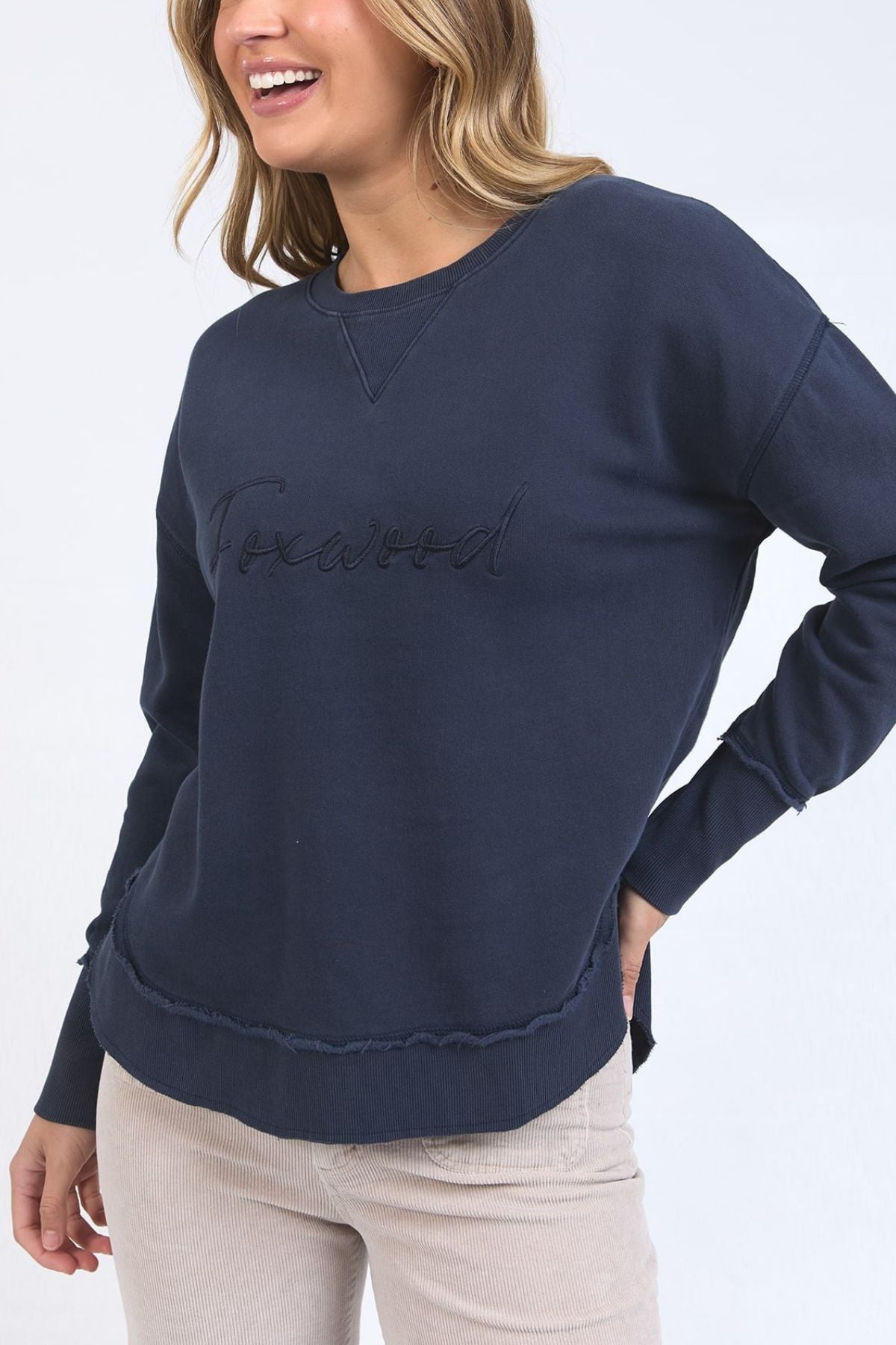 A woman smiles while wearing the Foxwood Signature Crew, a navy 100% cotton crewneck sweater with "Foxwood" embroidered on the front, paired with light beige pants against a plain light background.