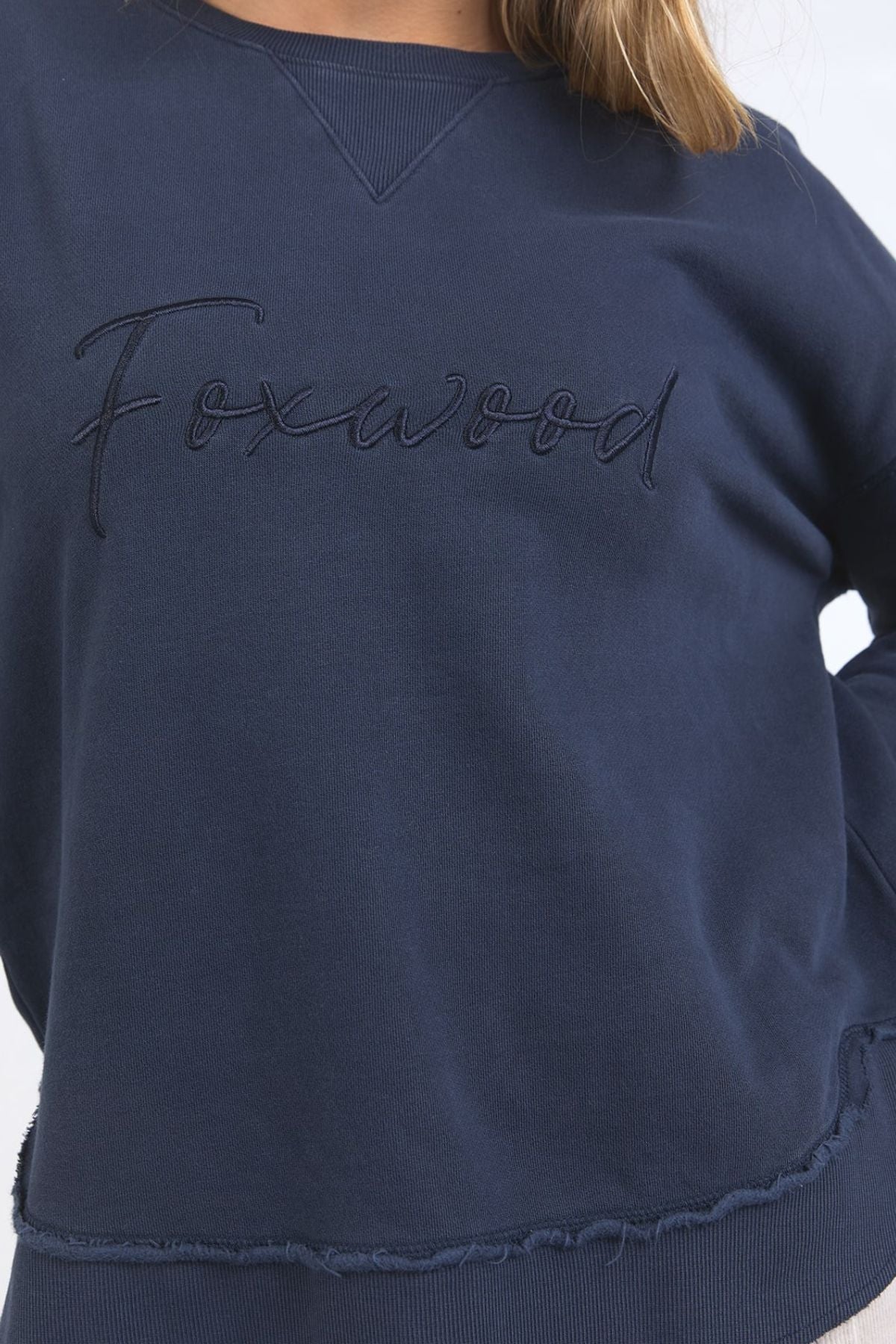 Someone wears the Foxwood Signature Crew, a navy crewneck sweater featuring "Foxwood" embroidered on the front in matching thread. Only their torso, part of an arm, and hair are visible.