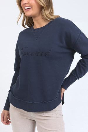 A woman smiles while wearing the Foxwood Signature Crew, a navy 100% cotton crewneck sweater with "Foxwood" embroidered on the front, paired with light beige pants against a plain light background.