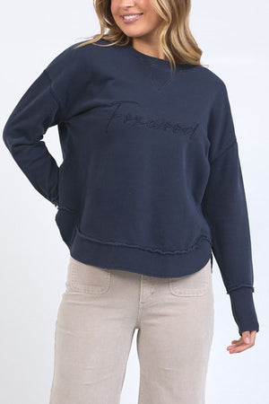A woman smiles with one hand on her hip, wearing the Foxwood Signature Crew— a navy blue 100% cotton crewneck sweater featuring "Foxwood" embroidery—paired with light beige pants.