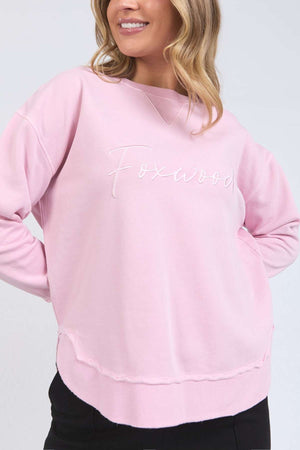 A woman smiles with her hands in her pockets, wearing the Foxwood Signature Crew—a relaxed-fit, light pink 100% cotton crewneck sweater featuring "Foxwood" embroidery and a rounded hem for effortless style.