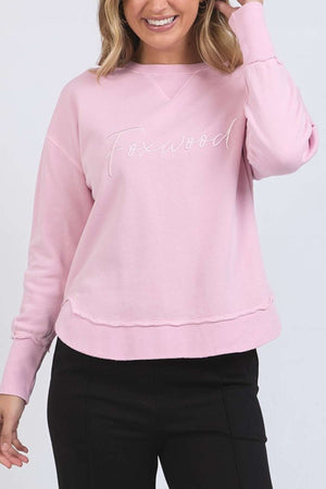 A woman smiles, touching her hair, wearing the light pink Foxwood Signature Crew—a 100% cotton crewneck sweater with "Foxwood" embroidered on the front—paired with black pants.