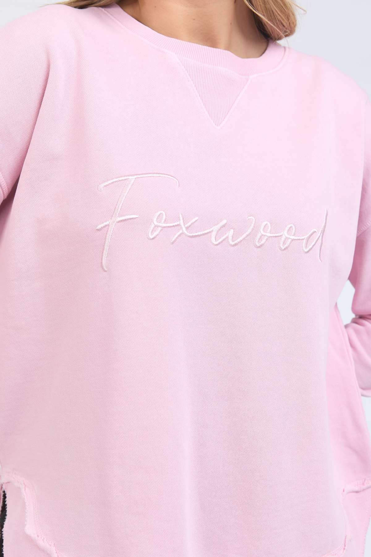 A person wears the Foxwood Signature Crew, a light pink crewneck sweater made of 100% cotton with "Foxwood" embroidered in white cursive on the chest. Only their upper torso and part of their face are shown.