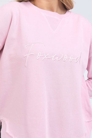 A person wears the Foxwood Signature Crew, a light pink crewneck sweater made of 100% cotton with "Foxwood" embroidered in white cursive on the chest. Only their upper torso and part of their face are shown.