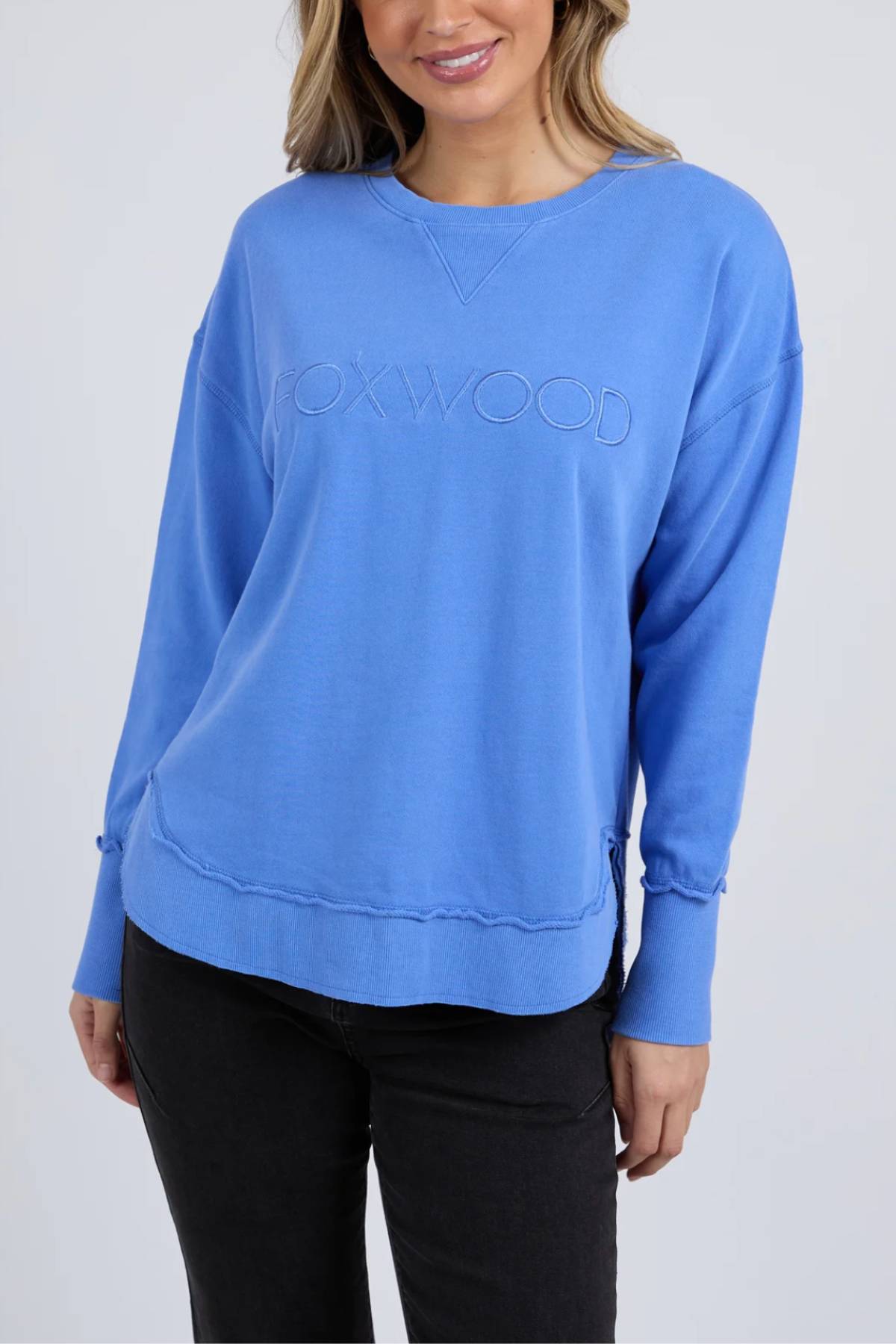 A woman smiles in loose waves, wearing a blue Foxwood Simplified Crew sweatshirt made from 100% cotton with "FOXYWOOD" embroidered on the front, paired with black jeans.