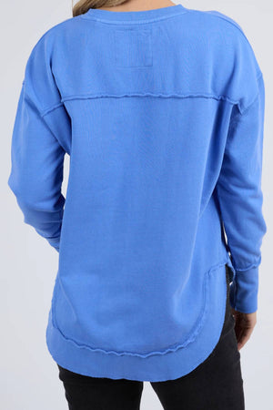 A person is seen from the back wearing a relaxed fit, long-sleeve blue Foxwood Simplified Crew sweatshirt with visible raw seams, 100% cotton fabric, and a rounded hem, paired with black pants.