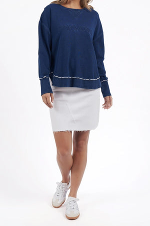 A woman stands against a white background wearing the navy blue Foxwood Simplified Crew, made from 100% cotton with "FOXWOOD" embroidered on the front, paired with a raw hem white skirt and white sneakers.