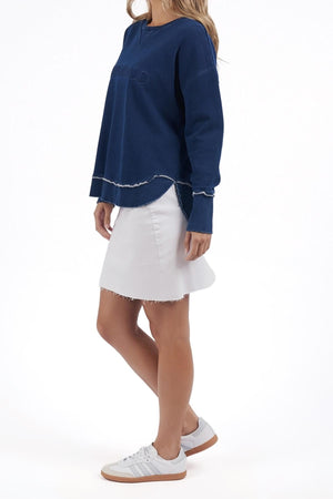 A woman wears the Foxwood Simplified Crew, a relaxed fit dark blue long-sleeve top with frayed edges, paired with a white skirt and sneakers, standing against a plain white background.