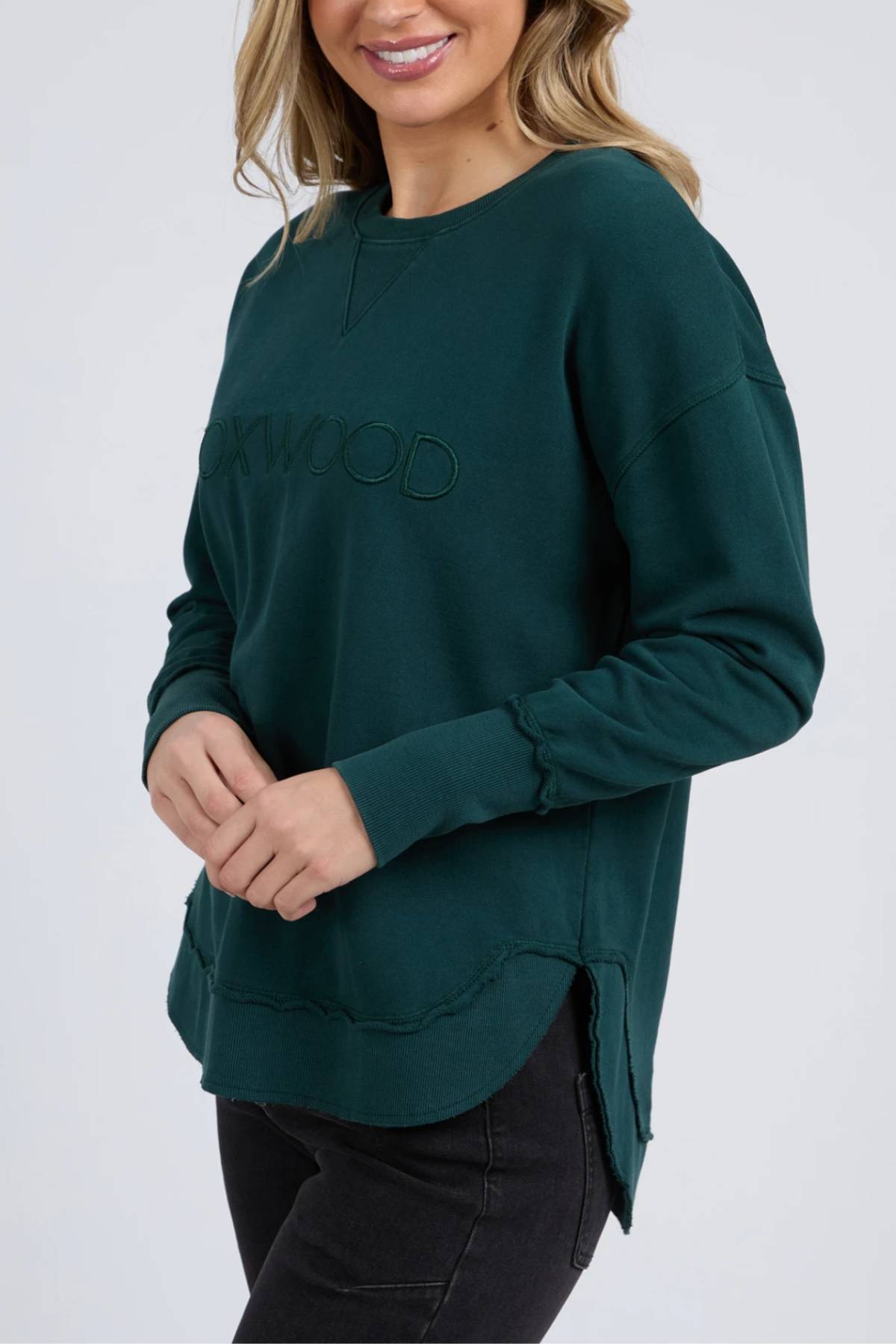 A woman wearing the Foxwood Simplified Crew, a dark green cotton sweatshirt with "ROOKWOOD" and Foxwood embroidery, stands with hands clasped in black pants against a light grey background.