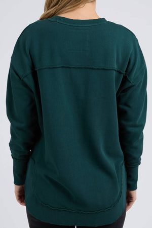 A person with long, light brown hair is shown from the back wearing the Foxwood Simplified Crew—a loose-fitting, dark teal 100% cotton sweatshirt with visible seams—paired with black pants against a plain light gray background.