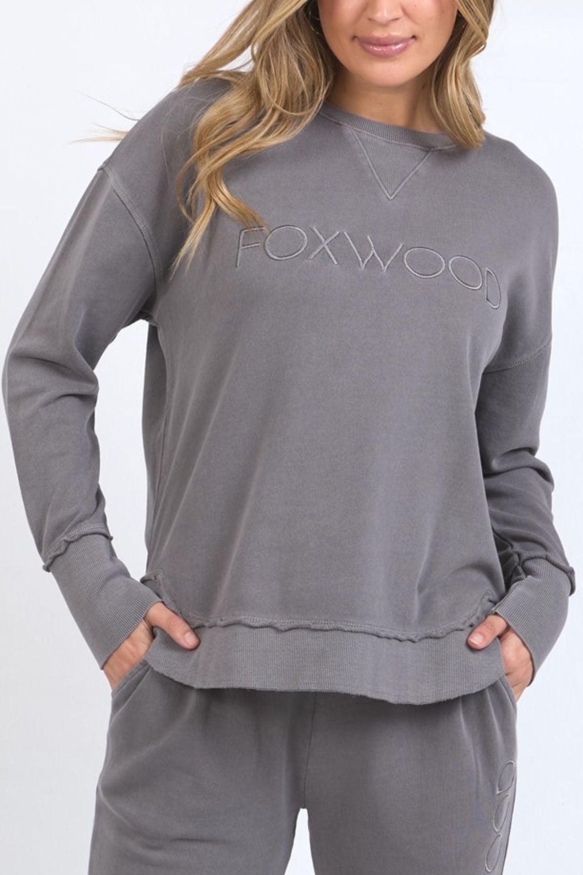 A woman with light wavy hair wears the loose-fit Foxwood Simplified Crew, a gray 100% cotton sweatshirt featuring front Foxwood embroidery, paired with matching sweatpants and her hands in her pockets.