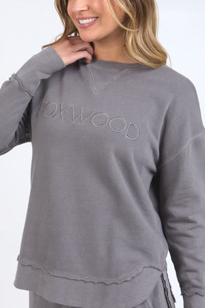 A woman in the Foxwood Simplified Crew, a relaxed-fit gray 100% cotton sweatshirt with Foxwood embroidery on the chest, smiles as she touches the collar with one hand.