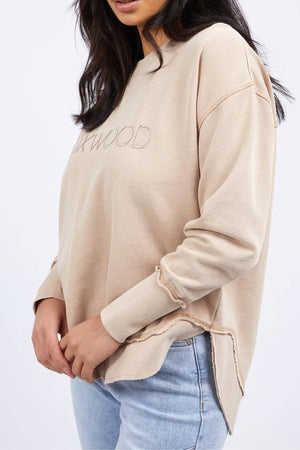 A woman with long dark hair wears a loose Foxwood Simplified Crew, featuring subtle embroidery, paired with light blue jeans. She stands against a plain white background, head slightly turned and hands gently clasped in front.