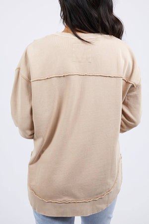 A person with long dark hair is shown from behind, wearing a beige Foxwood Simplified Crew made from 100% cotton with exposed seams, paired with light blue jeans against a plain, light background.