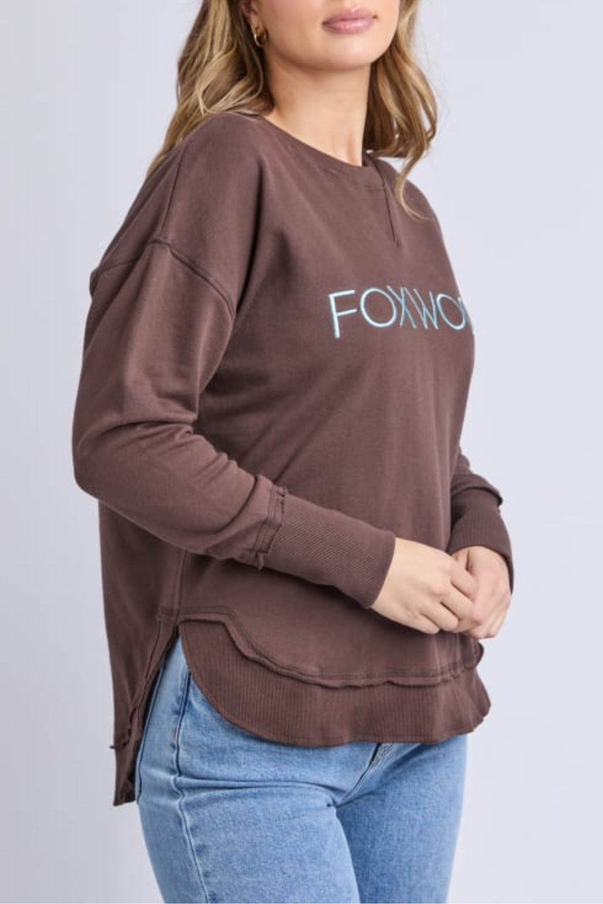 A woman in a loose-fitting brown Foxwood Simplified Metallic Crew sweatshirt with a light blue logo and light blue jeans stands against a plain light gray background.