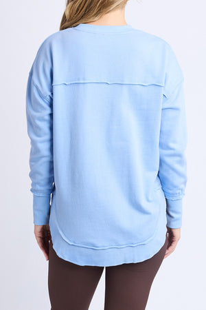 A person with long, light hair is seen from the back wearing the loose-fitting Foxwood Simplified Metallic Crew women's sweatshirt with lurex thread, a horizontal seam, ribbed cuffs, and dark brown pants.
