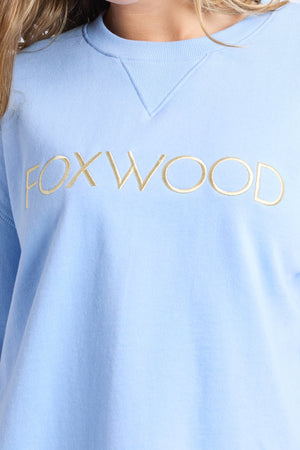 Close-up of a person wearing the Foxwood Simplified Metallic Crew, a women's sweatshirt featuring "FOXWOOD" embroidered in gold lurex thread across the chest.