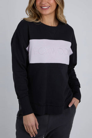 A woman models the Foxwood Simplified Tonal Crew, a black cotton fleece with a light pink panel and "FOXWOOD" on the chest. She smiles with one hand in her pocket, highlighting the stylish hi-lo hemline against a plain background.