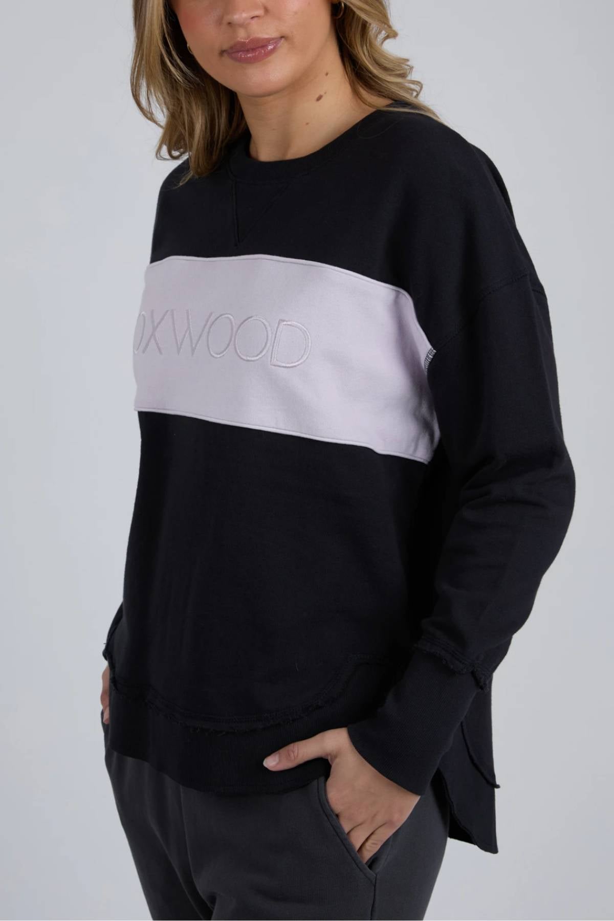 A woman with light brown, wavy hair wears the Foxwood Simplified Tonal Crew, a black cotton fleece crew with a light gray horizontal panel and embroidered text, paired with dark pants and one hand in her pocket.