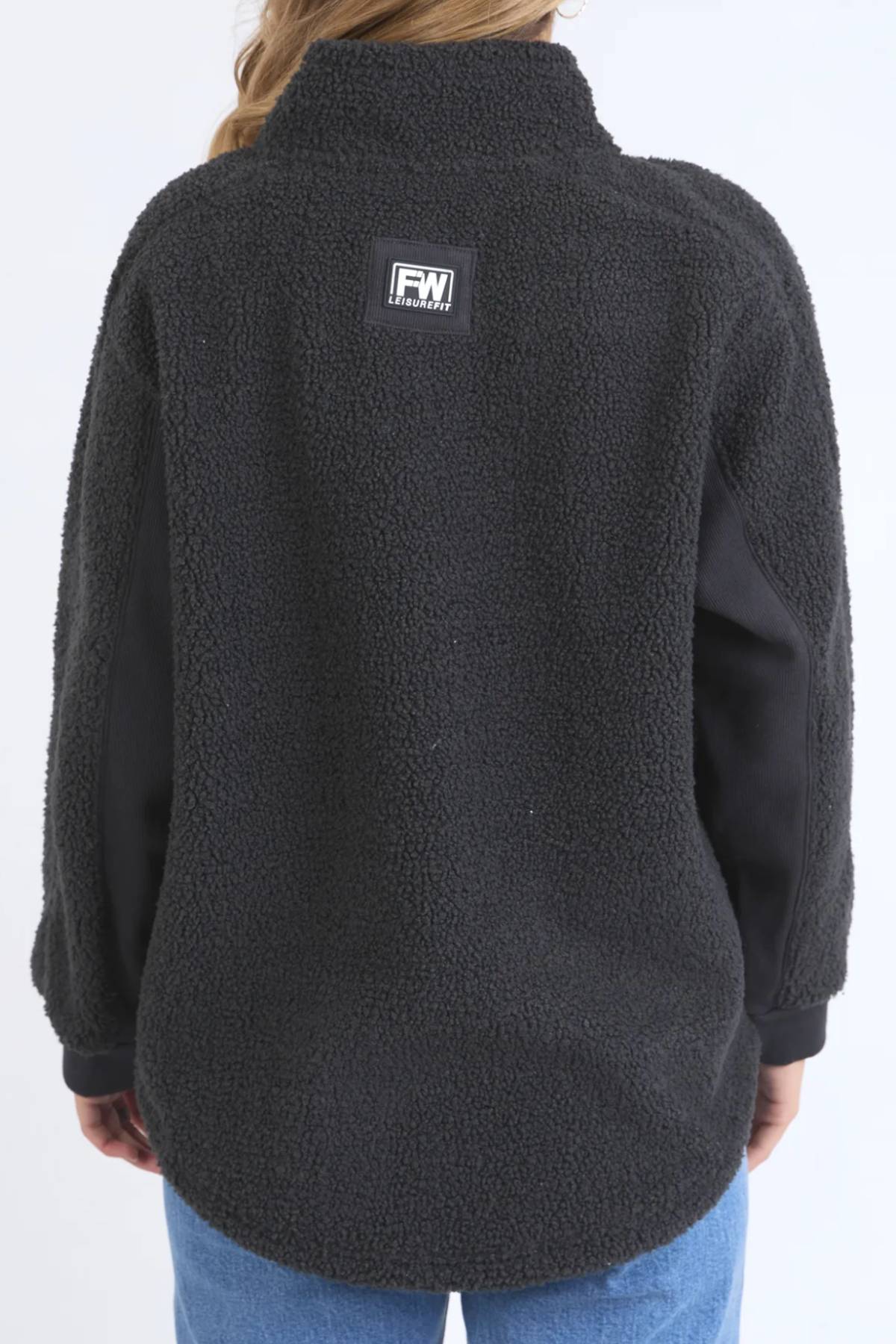 A person wears the black Foxwood Weekend Boucle Crew, featuring a textured finish and an FW logo patch on the upper back, styled with light blue jeans—ideal cozy loungewear for relaxing days—against a plain light background.