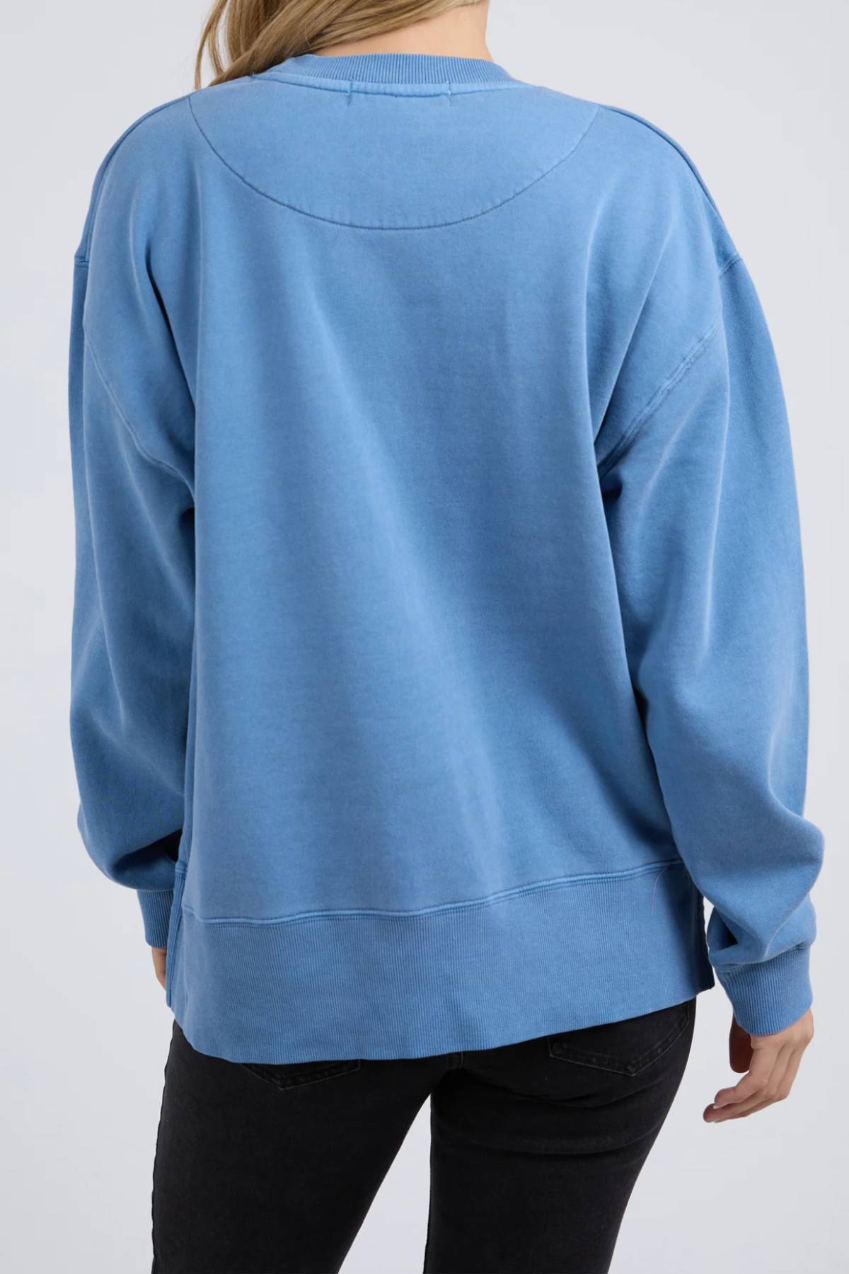 A person with long blonde hair is seen from the back in a light blue Foxwood Trademark Crew and black pants. The scene has a simple, light background, highlighting this crew as a versatile staple for any casual wardrobe.