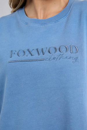 A person wears the Foxwood Trademark Crew, a blue unbrushed cotton fleece sweatshirt with "FOXWOOD clothing" embroidered on the chest. Only their lower face, neck, and upper torso are visible—perfect for any casual wardrobe.