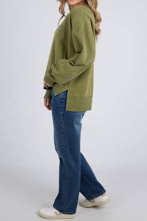 Dressed in a loose olive green Foxwood Trademark Crew, blue jeans, and white sneakers, a person stands sideways against a plain background—an ideal way to style the versatile Foxwood Trademark Crew for any casual occasion. Face is not visible.