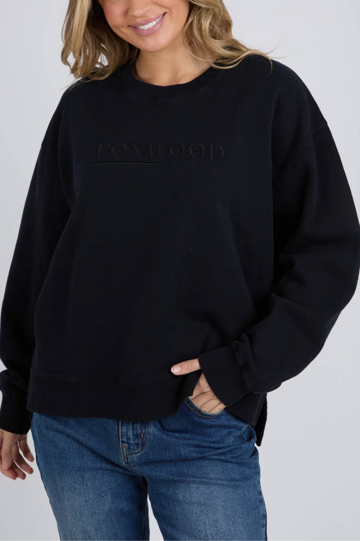 Wearing the Foxwood Trademark Crew, a relaxed-fit black sweatshirt in unbrushed cotton fleece, she pairs it with blue jeans and smiles with one hand in her pocket—an ideal piece for any casual wardrobe.