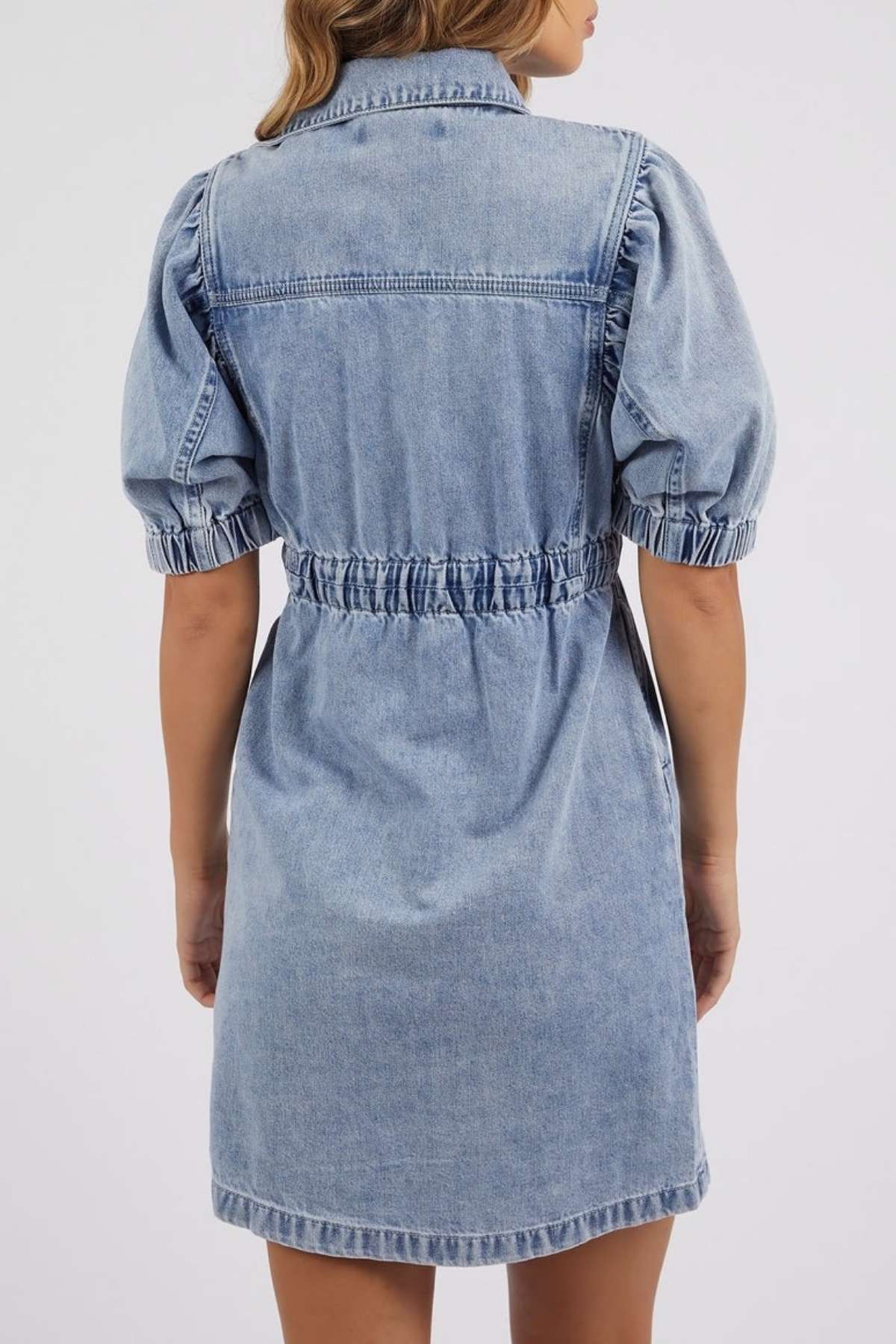 A woman is shown from behind wearing the Foxwood Vivian Dress, a light blue stretch denim dress with short puffed sleeves, an elastic waist, and visible stitching.