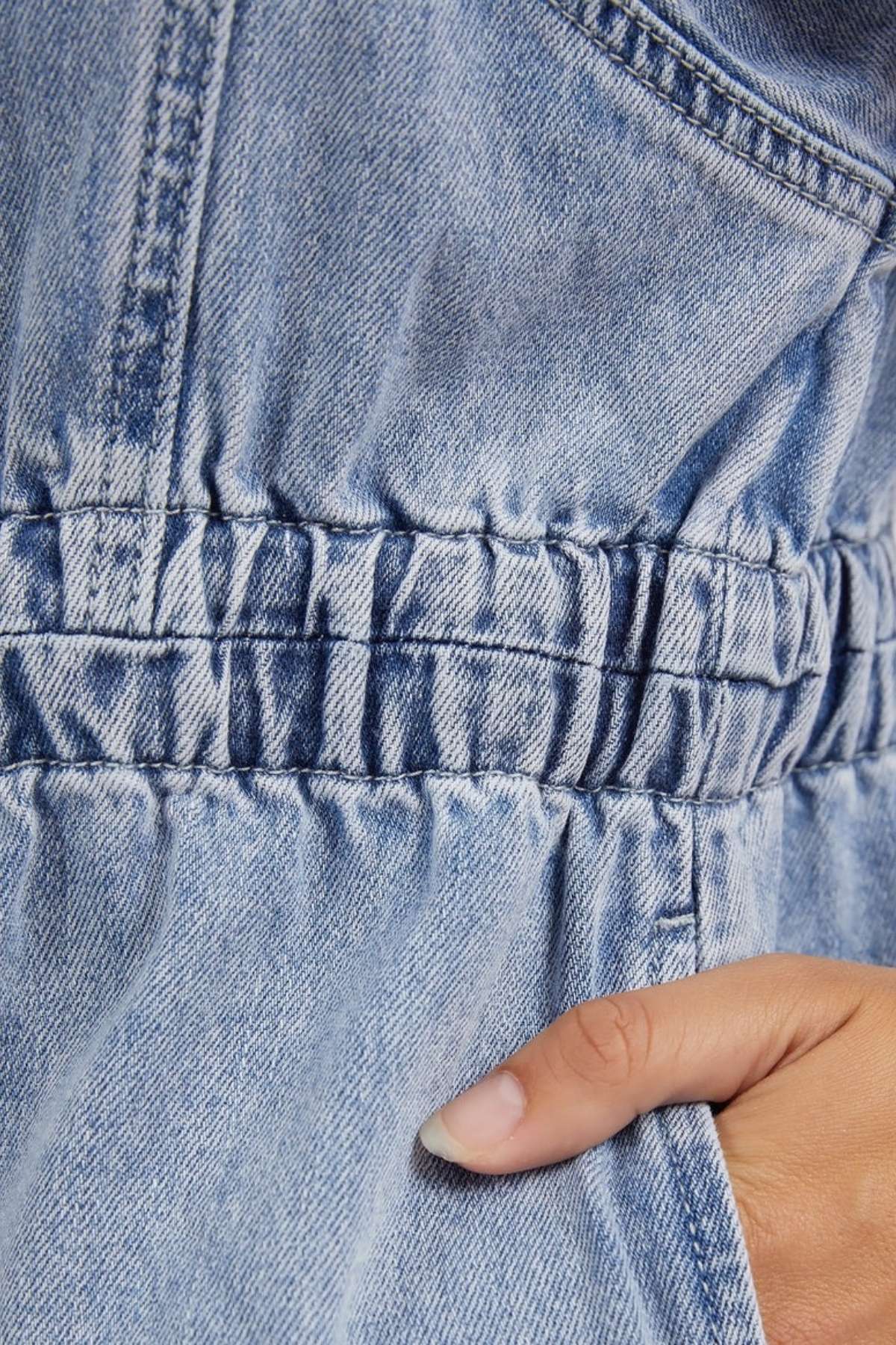 A close-up of the Foxwood Vivian Dress, featuring light blue stretch denim with gathered stitching at the elastic waist and a hand with light-colored nails tucked into a side pocket.