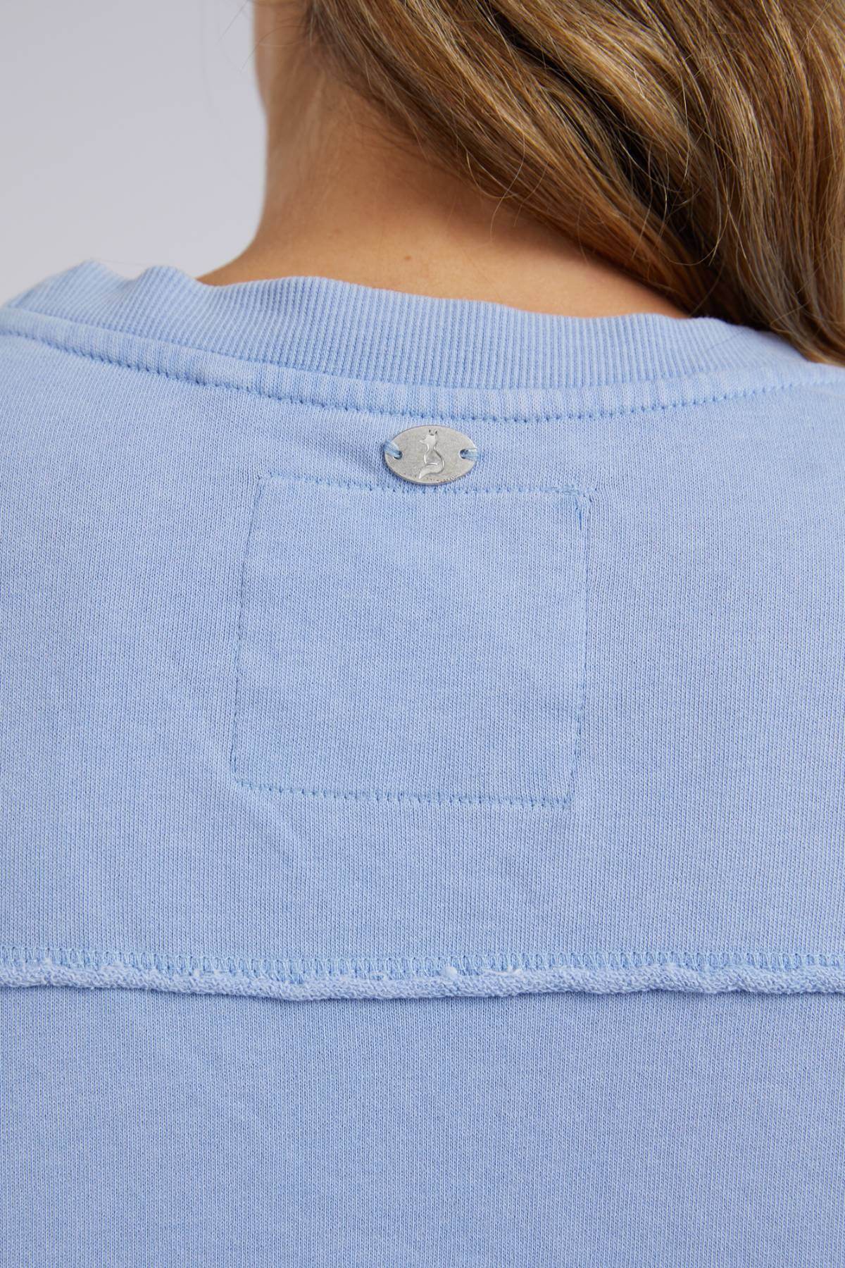 Close-up of a person wearing a comfortable Foxwood Washed Simplified Crew in light blue, made of 100% cotton. The shirt has a visible seam and features a small silver logo near the collar, partially obscured by the person's hair.