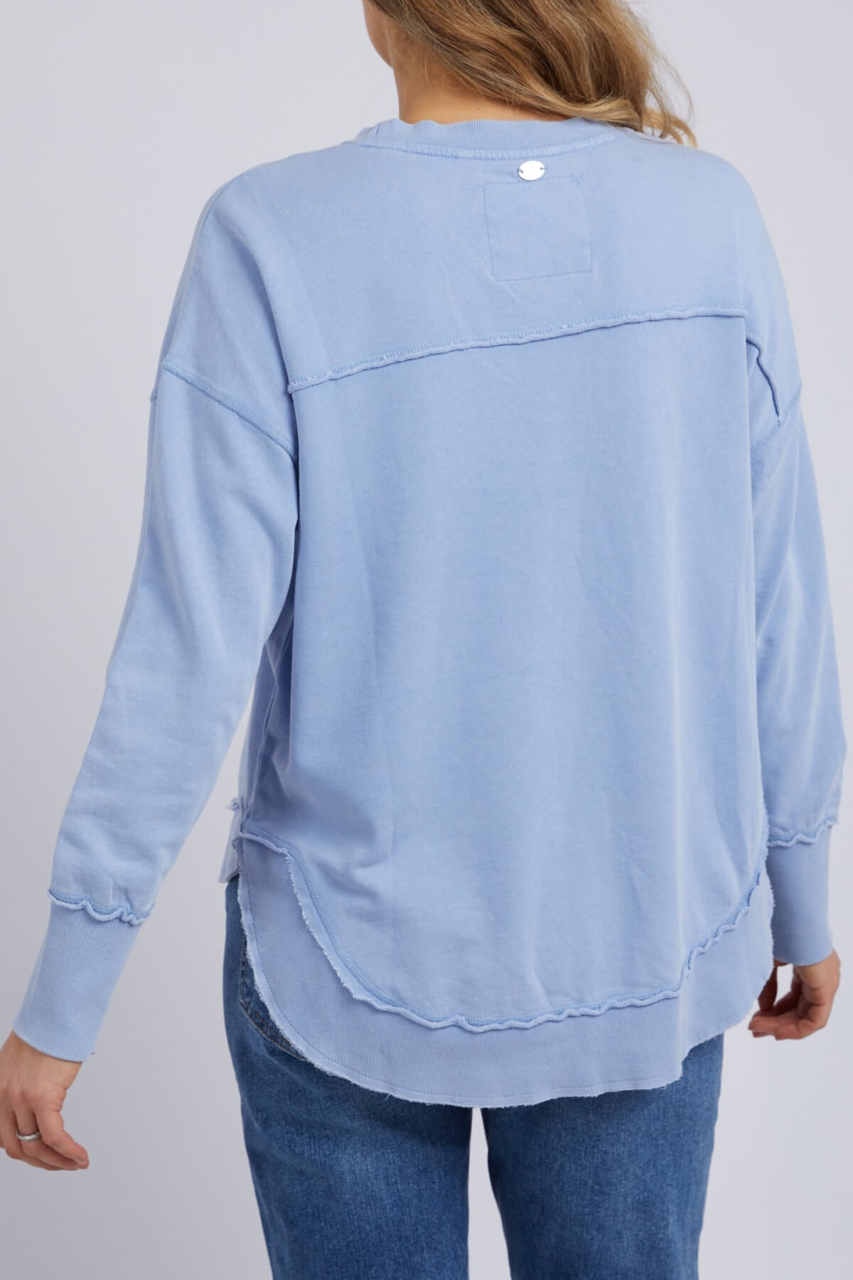 Seen from behind, the individual with long hair wears the Foxwood Washed Simplified Crew— a light blue, 100% cotton long-sleeve shirt featuring decorative seams and a rounded hem—paired with blue jeans against a plain light gray background.