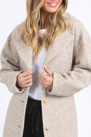 A woman with wavy blonde hair smiles, holding closed her light beige Foxwood Whistler Coat over a white top and black pants. The wool-blend fashion coat is partially concealing her face.
