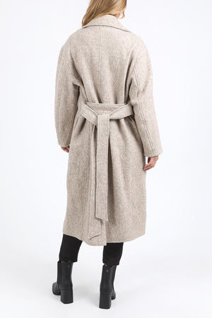 Wearing the Foxwood Whistler Coat—a long, light beige coat—a person in black pants and ankle boots stands with their back to the camera against a plain white background.
