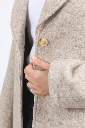 Wearing the Foxwood Whistler Coat, a fashion piece, a person closes it with one hand, emphasizing the large tan button while showcasing the coat’s texture and their gesture.
