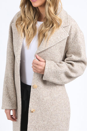 A woman with long, wavy blonde hair wears the Foxwood Whistler Coat—light beige and stylish—holding the lapel as she stands against a plain background, her face partly out of frame.