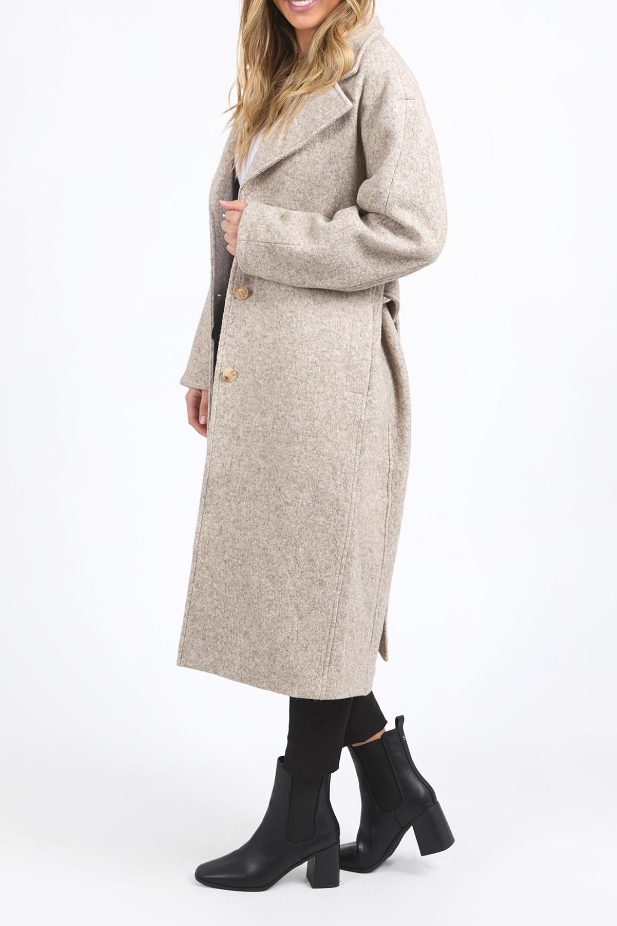 A woman models the long beige Foxwood Whistler Coat with large lapels, styled with black leggings and heeled ankle boots, standing against a plain white background. Her face is partially out of frame.