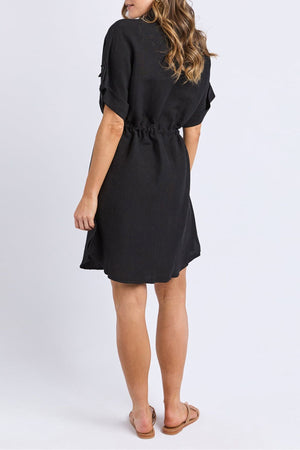 A woman faces away, wearing a short-sleeved black Foxwood Willow Shirt Dress with a cinched waist and brown sandals. Her wavy, light brown hair falls over her shoulders against a plain white background.