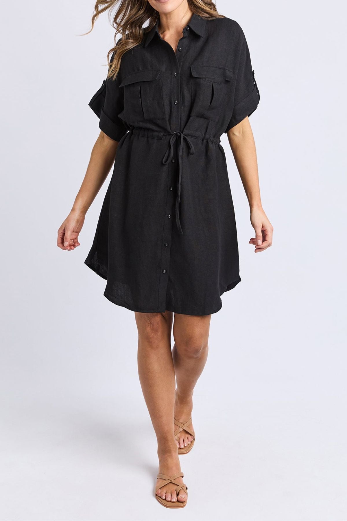 A woman wears the Foxwood Willow Shirt Dress, a short-sleeve black dress with a drawstring waist and chest pockets, styled with flat sandals against a plain light background.