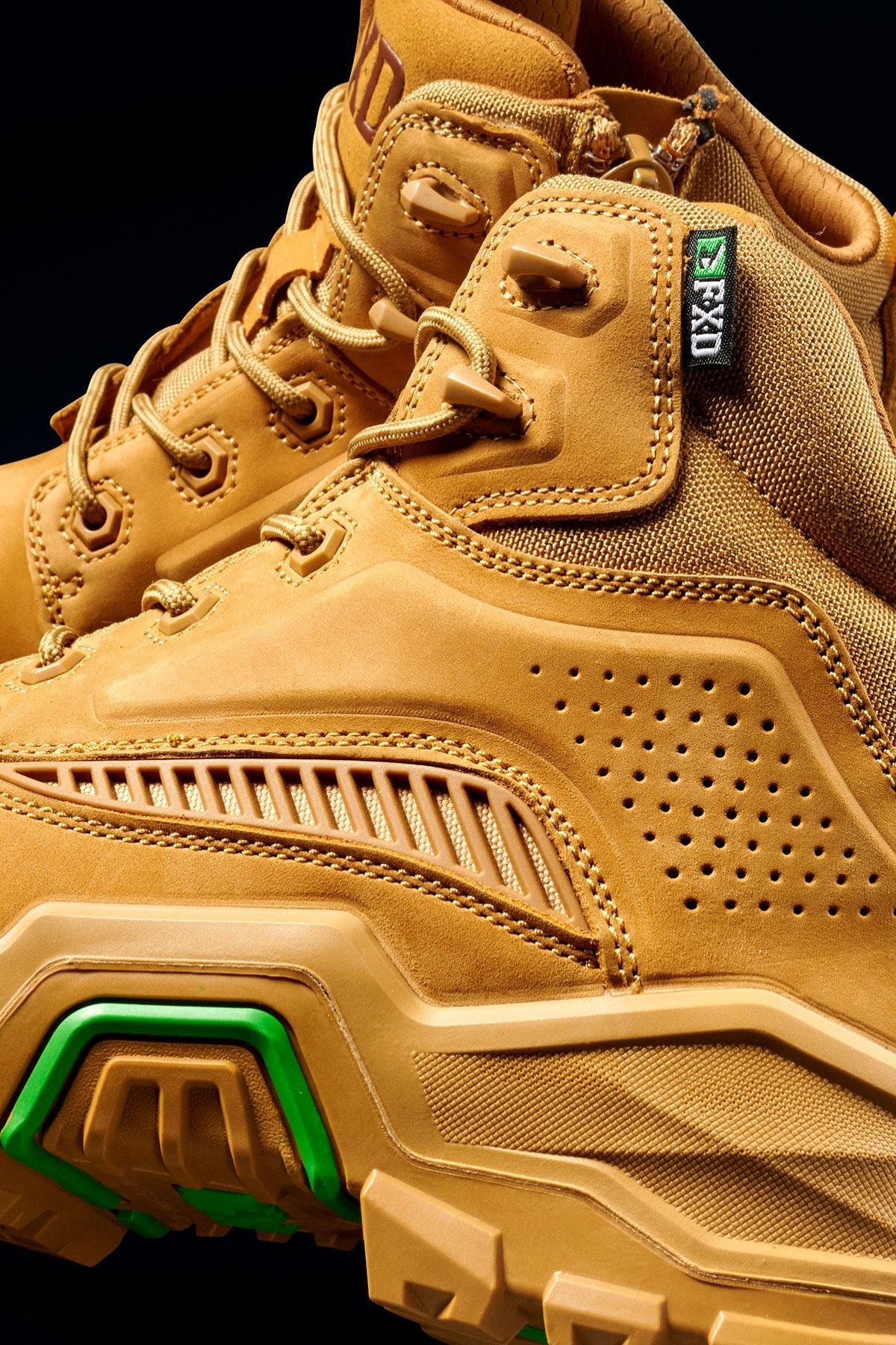 Close-up of FXD Dura X-2 Nitrocore™ Mid Cut High Performance Work Boots featuring tan textured leather, green sole accents, detailed stitching, perforated side panels, and sturdy laces against a black background.