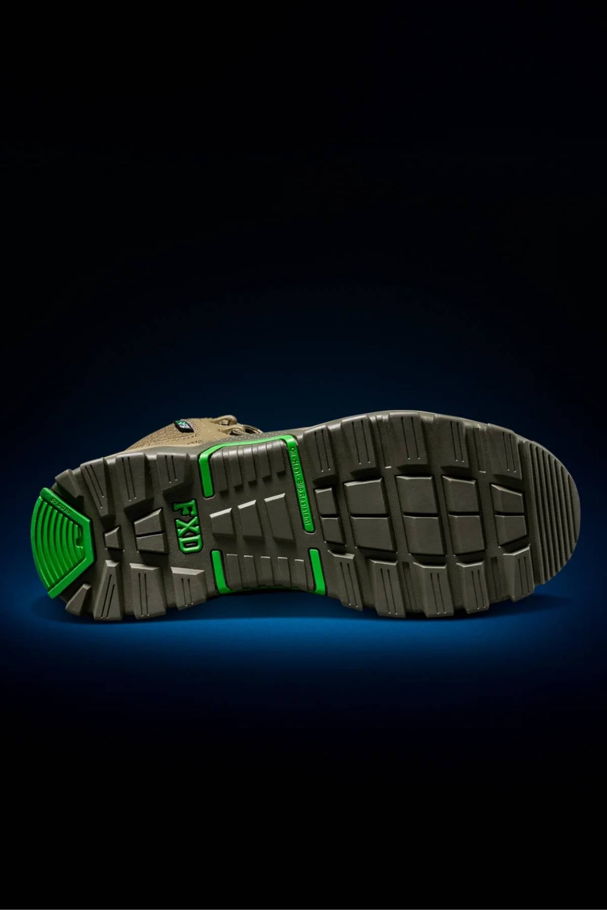 The image displays the sole of the FXD WB-6 Work Boot, featuring a rugged brown design with bright green accents. Its deep treads and textured pattern ensure superior grip and meet tough safety standards, shown against a dark background.